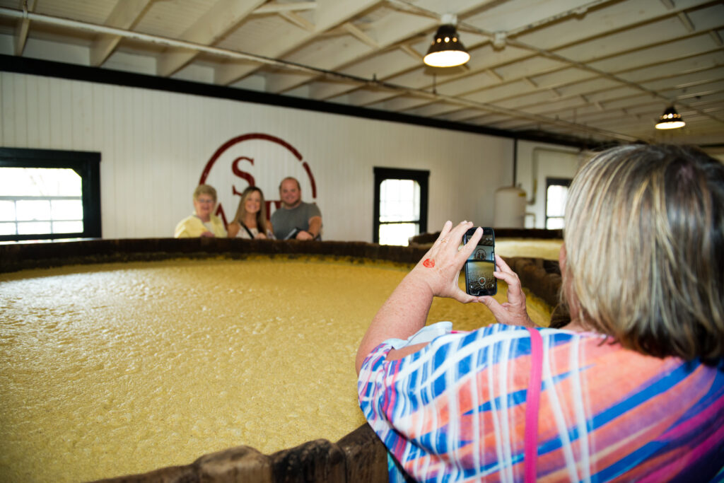 Tourist taking a photo of her friends in front of a giant vat of fermenting bourbon while on tour at Makers Mark in Loretto, KY